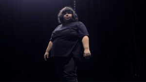 Jussara Belchior, fat dancer, dressed in black in a black space, looking down at the camera.